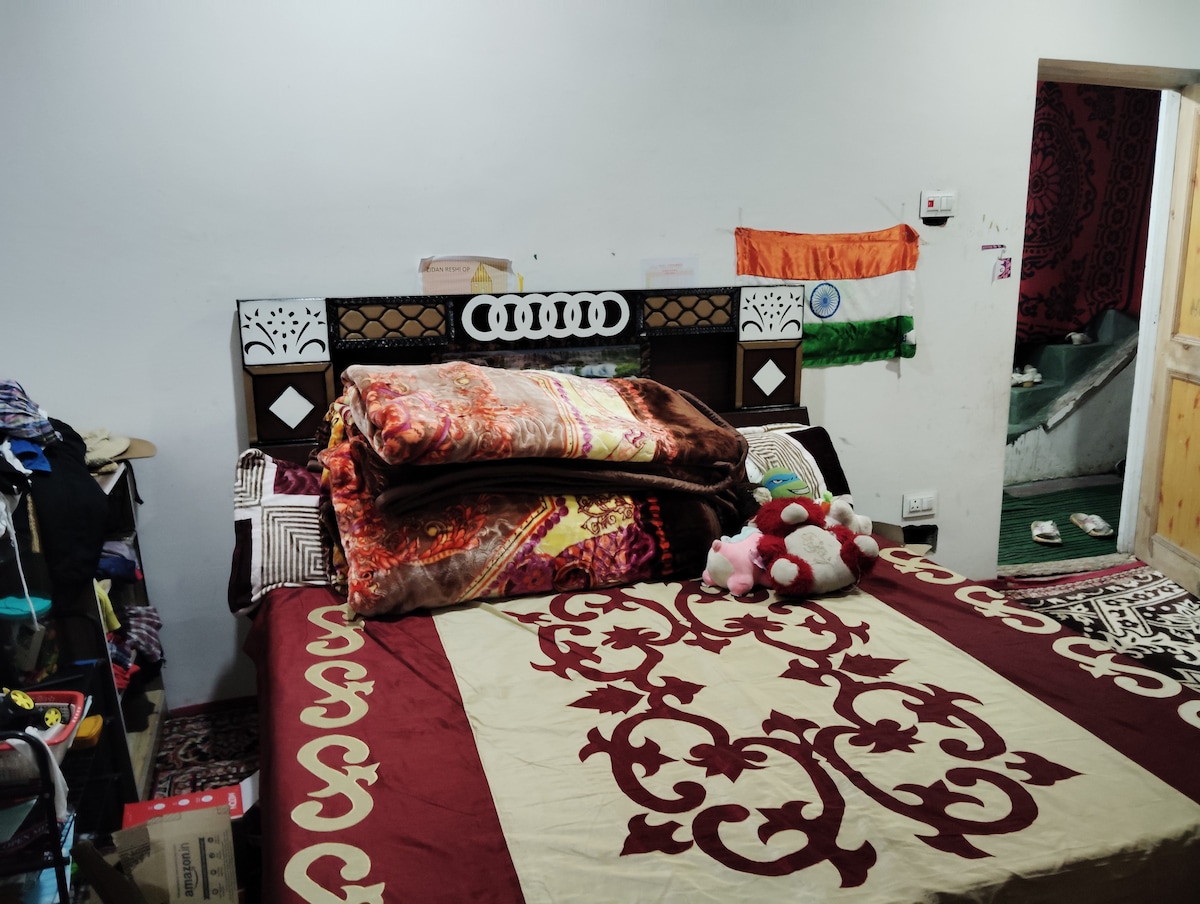 A cozy bedroom features a large bed adorned with layered, colorful blankets. A decorative headboard adds detail to the space. Nearby, shelves are filled with clothing, and an Indian flag is displayed on the wall, providing a personal touch to the room.