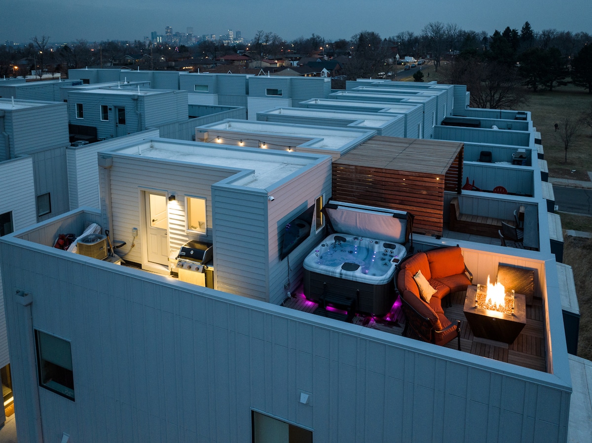 The rooftop patio features a premium hot tub, seating area with orange cushions, and a fire pit, all illuminated with soft lighting. The kitchen area is visible, highlighting convenience for outdoor dining. The city skyline is faintly seen in the background.