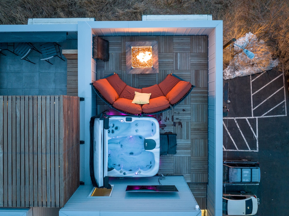 The rooftop patio features a premium hot tub accompanied by a fire pit and seating area. A cozy orange loveseat and a decorative pillow are positioned near the fire, offering a space for relaxation. A partial view of the surrounding area is visible.