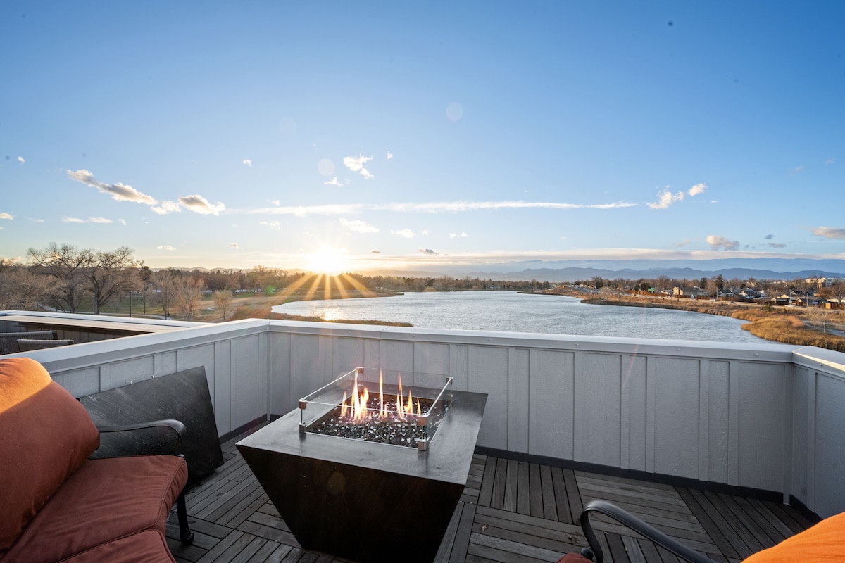 A rooftop patio is presented, featuring a fire pit surrounded by seating. The sun is setting over Rocky Mountain Lake, casting rays of light across the water. Expansive views of the landscape are visible, enhancing the outdoor experience.