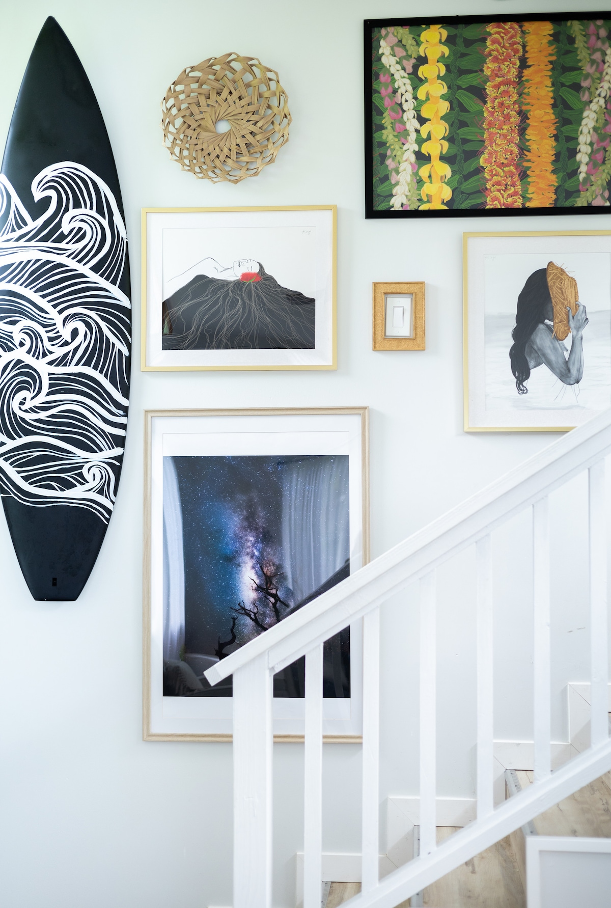 A collection of framed artworks is displayed on a light-colored wall, featuring a black surfboard alongside vibrant prints. A large photograph of a starry night sky is positioned centrally, with various other art pieces surrounding it, all enhancing the space's artistic ambiance.