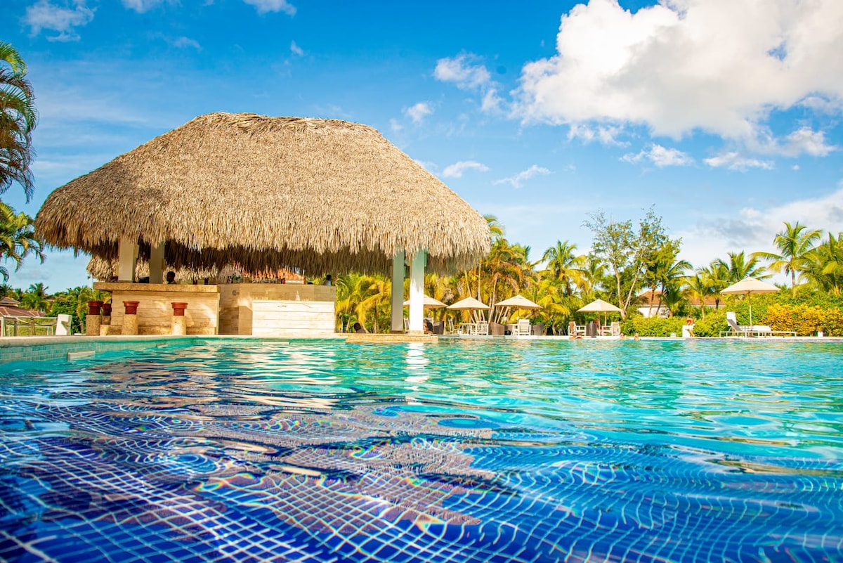 A tranquil swimming pool glistens under clear skies, featuring an inviting palapa-style bar at the water's edge. Sun loungers are positioned nearby, surrounded by lush palm trees and vibrant greenery, offering a serene environment for relaxation.