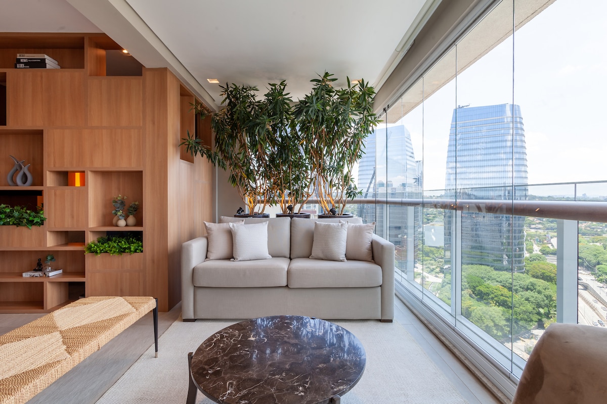 A modern living area features a light-colored sofa adorned with plush pillows, positioned beside a coffee table with a dark tabletop. Lush indoor plants are displayed prominently, and large glass windows offer a view of the surrounding buildings and green landscape.