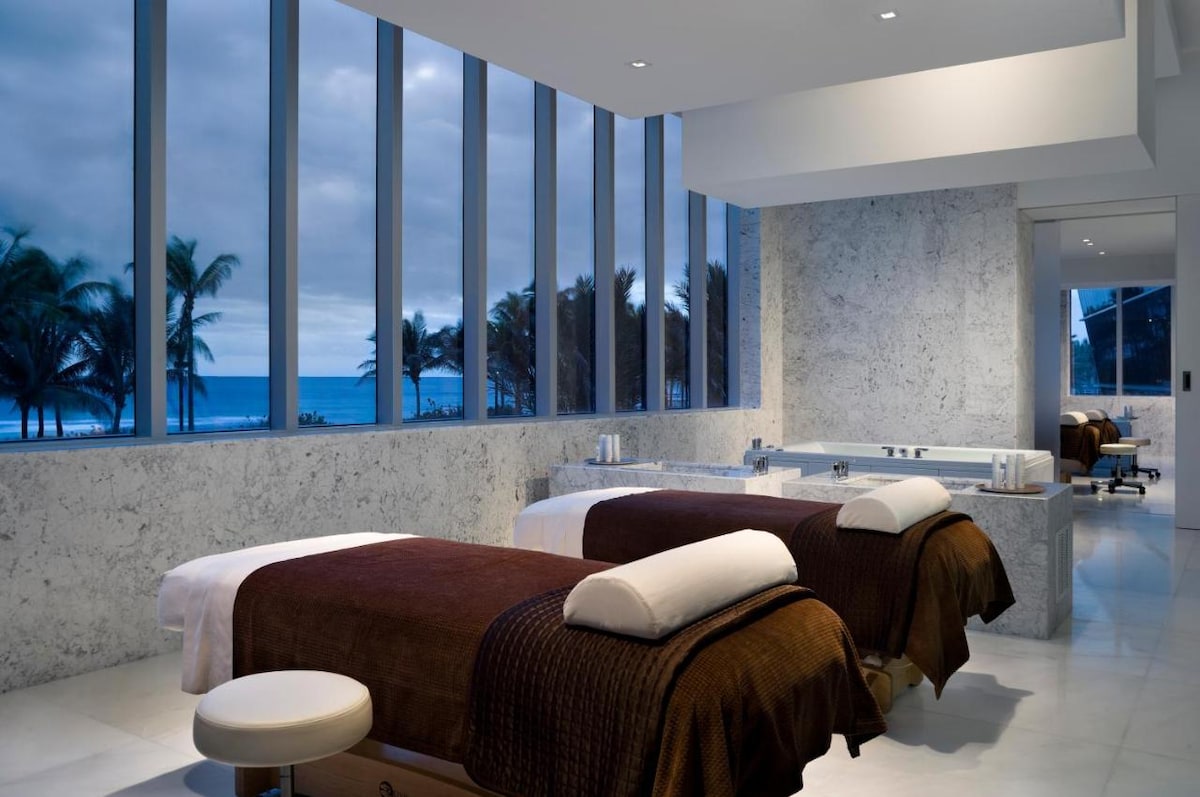 A serene spa room features two treatment beds positioned beside expansive floor-to-ceiling windows, offering views of palm trees and the ocean. The room is adorned with marble finishes, creating a tranquil atmosphere. Soft lighting and a neutral color palette contribute to the calming environment.