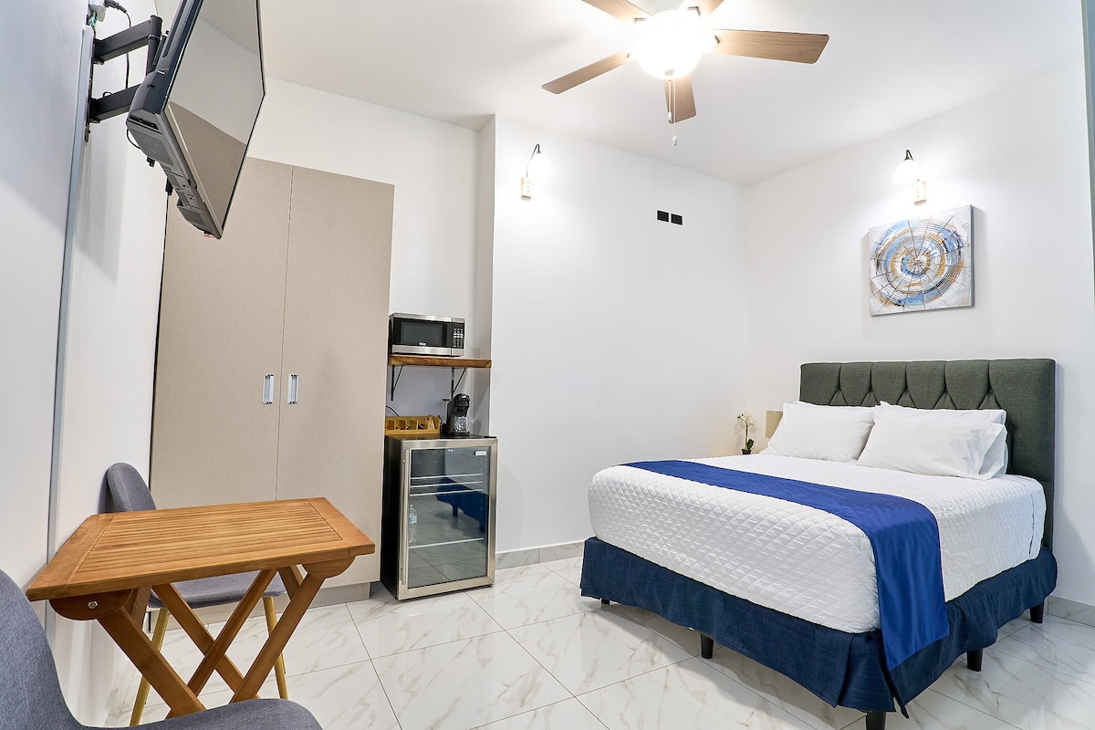 A well-appointed room is featured, showcasing a spacious bed with crisp linens. A mounted television and a small wooden table are prominently displayed, while a microwave and mini-fridge are positioned on a shelving unit. Natural light illuminates the room through the nearby wall art.