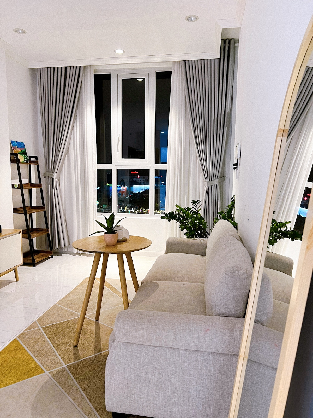 A cozy seating area features a light-colored sofa paired with a round wooden table. Plants are positioned near the window, which is framed by long, light gray curtains. A mirror is seen reflecting the space, adding depth to the room.