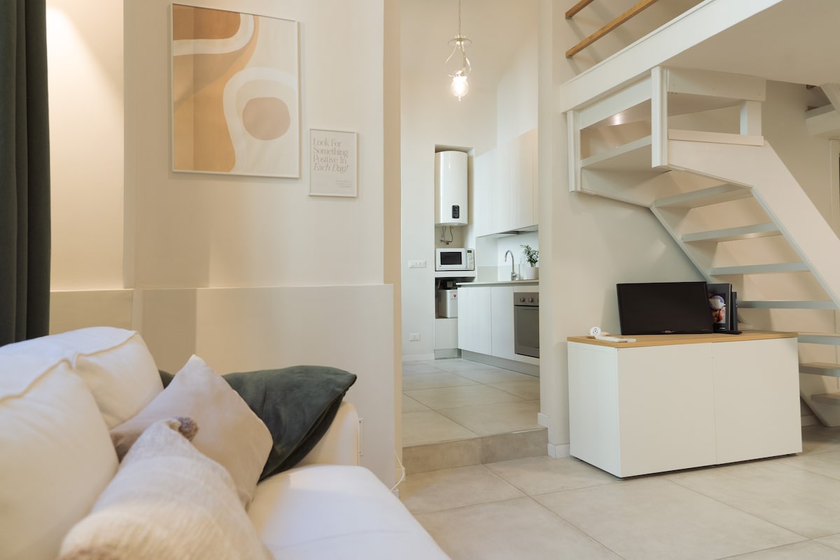 A spacious living area features a white sofa adorned with cushions, complemented by an abstract art piece on the wall. The open layout leads to a modern kitchen area equipped with essential appliances, visible in the background. Light fills the space, enhancing its functional design.