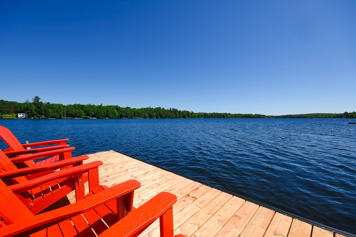 A wooden dock extends over a serene lake, featuring several bright red Adirondack chairs positioned for relaxation. The calm water reflects the clear blue sky, bordered by lush greenery on the opposite shore, creating an inviting lakeside atmosphere.