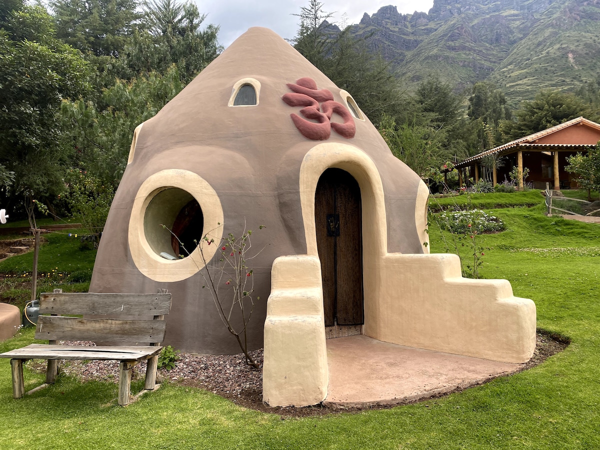 A charming dome-shaped structure is set within a lush green garden, featuring multiple round windows and a decorative motif above the entrance. The natural materials used create a harmonious blend with the surrounding landscape, while a wooden bench is placed nearby.