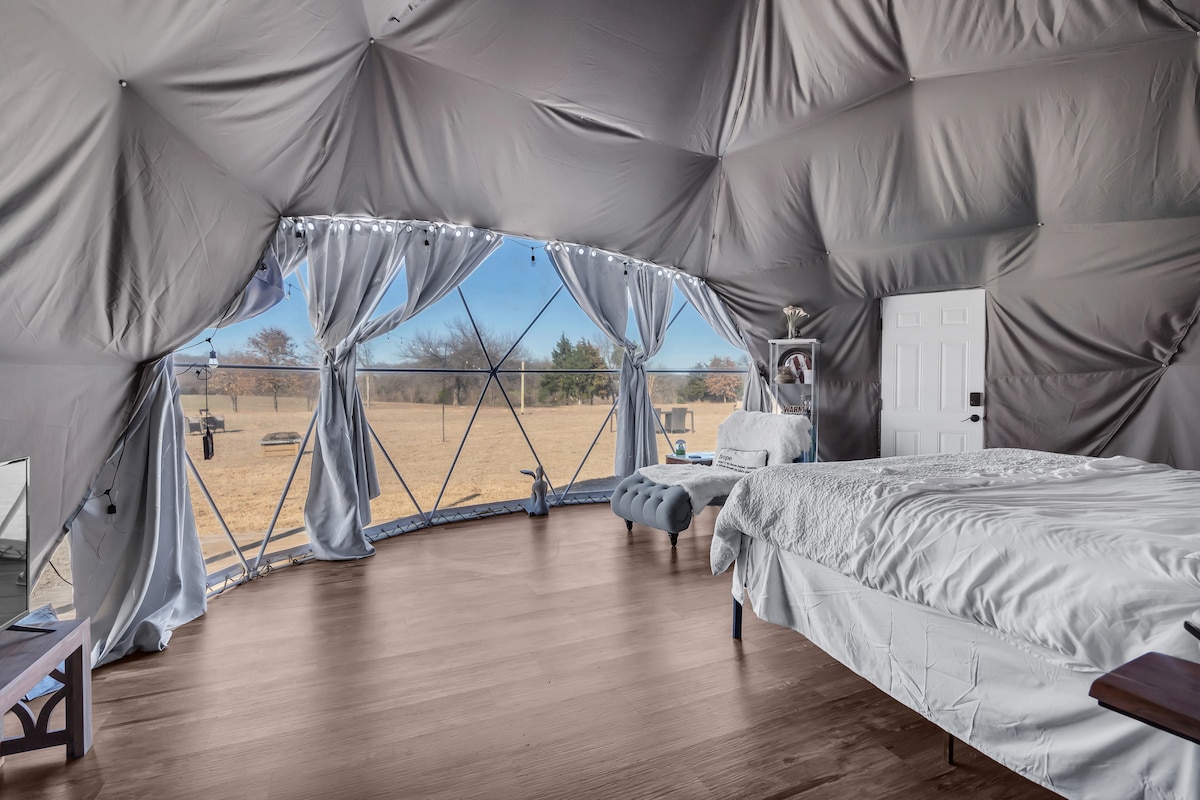 Inside the event dome, a spacious room is highlighted by large windows allowing natural light and views of the grassy landscape. A king bed is positioned to one side, complemented by a cozy seating area. Soft curtains frame the windows, enhancing the serene ambiance.