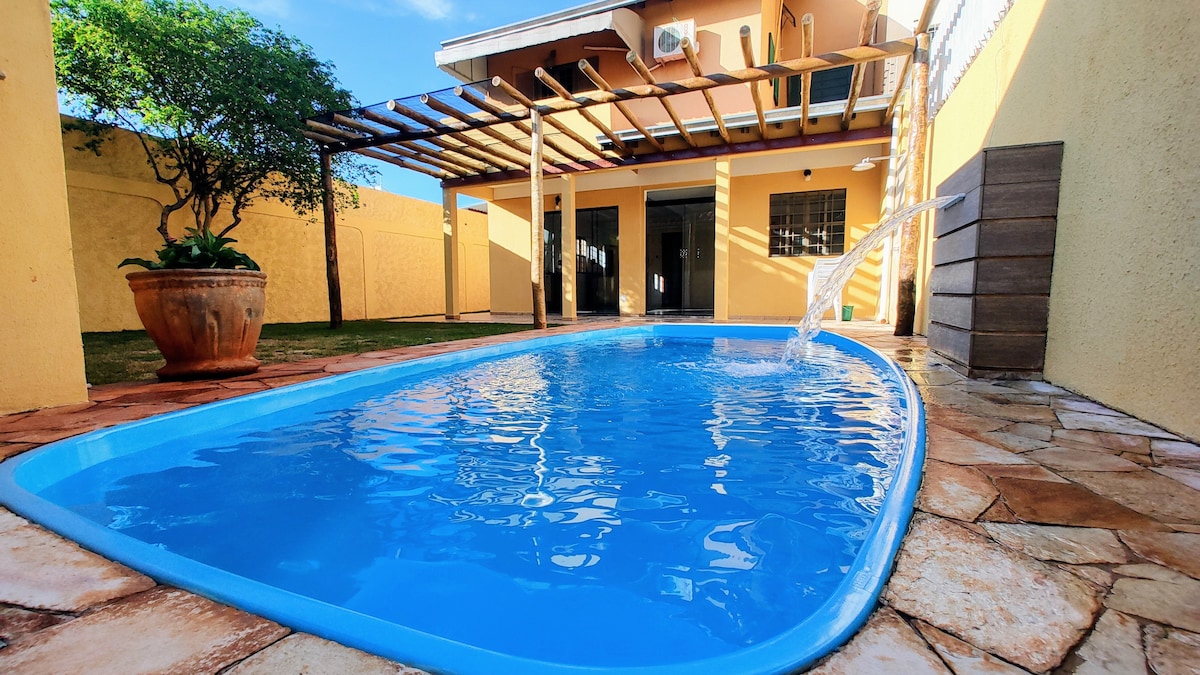 A clear blue swimming pool is visible, with water gently flowing from a fountain. Surrounding the pool, a spacious yard is bordered by a yellow wall and features a wooden pergola overhead. Sunlight brightens the inviting outdoor space.