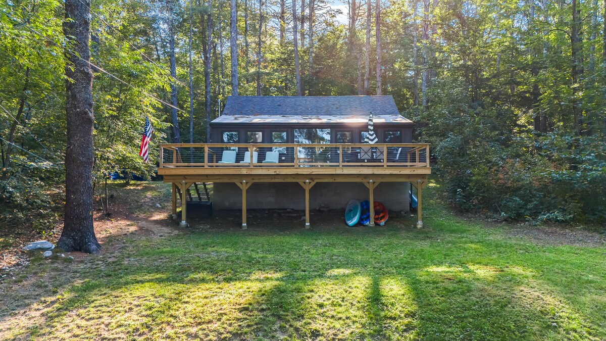 A modern cabin is positioned on a grassy area surrounded by tall trees, featuring a spacious deck. The structure has large windows that invite natural light, while colorful kayaks and paddle boards are stored nearby, hinting at outdoor activities available for guests.