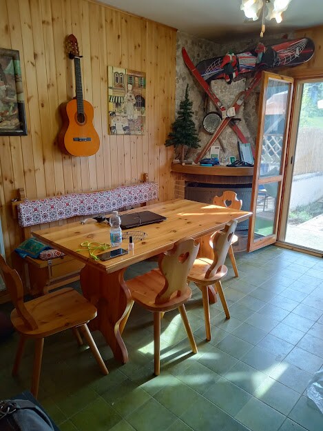 The dining area features a wooden table surrounded by four chairs, with a small bench along the wall. Decor includes a guitar hanging on wooden paneling and ski equipment displayed nearby. Natural light enters through a glass door leading to the exterior space.