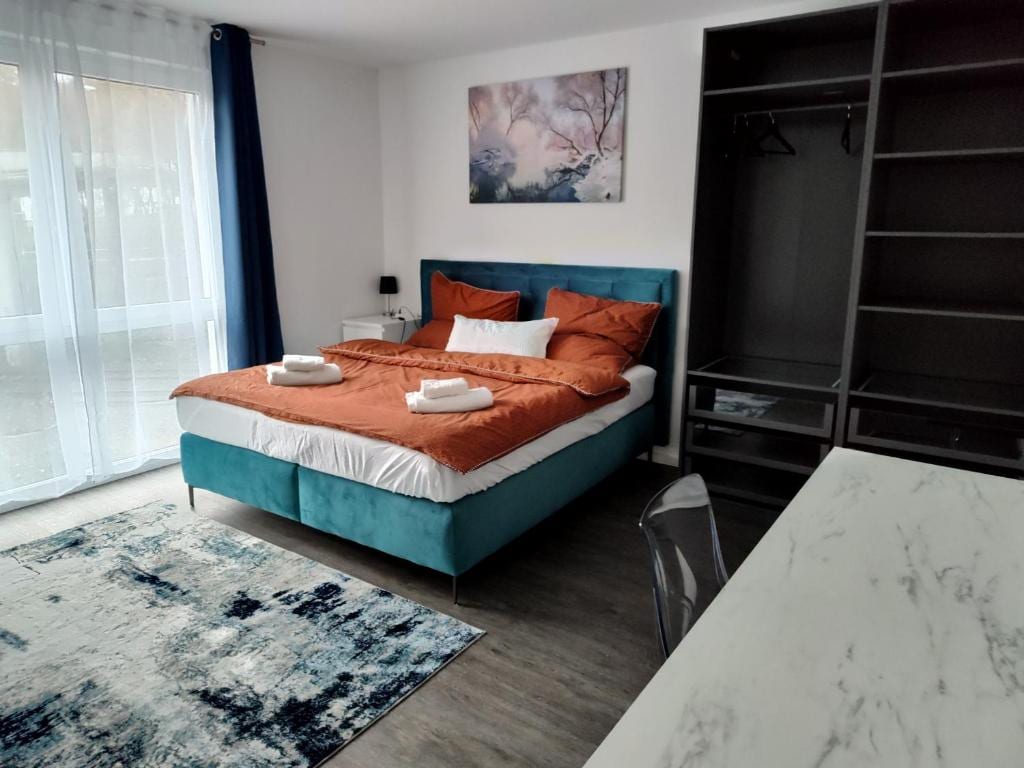 A well-appointed sleeping area features a spacious bed dressed in warm orange linens, accompanied by neatly arranged towels. A light-toned rug adds texture to the floor, while large windows allow natural light to brighten the room. A modern wardrobe and desk enhance the functional space.