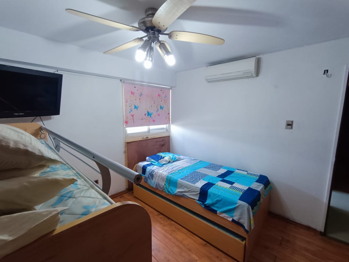 A well-lit bedroom features two beds, one elevated with a built-in design and the other positioned below. A ceiling fan with multiple lights provides illumination, and a wall-mounted television is visible. Colorful bedding adds a touch of vibrancy to the space.