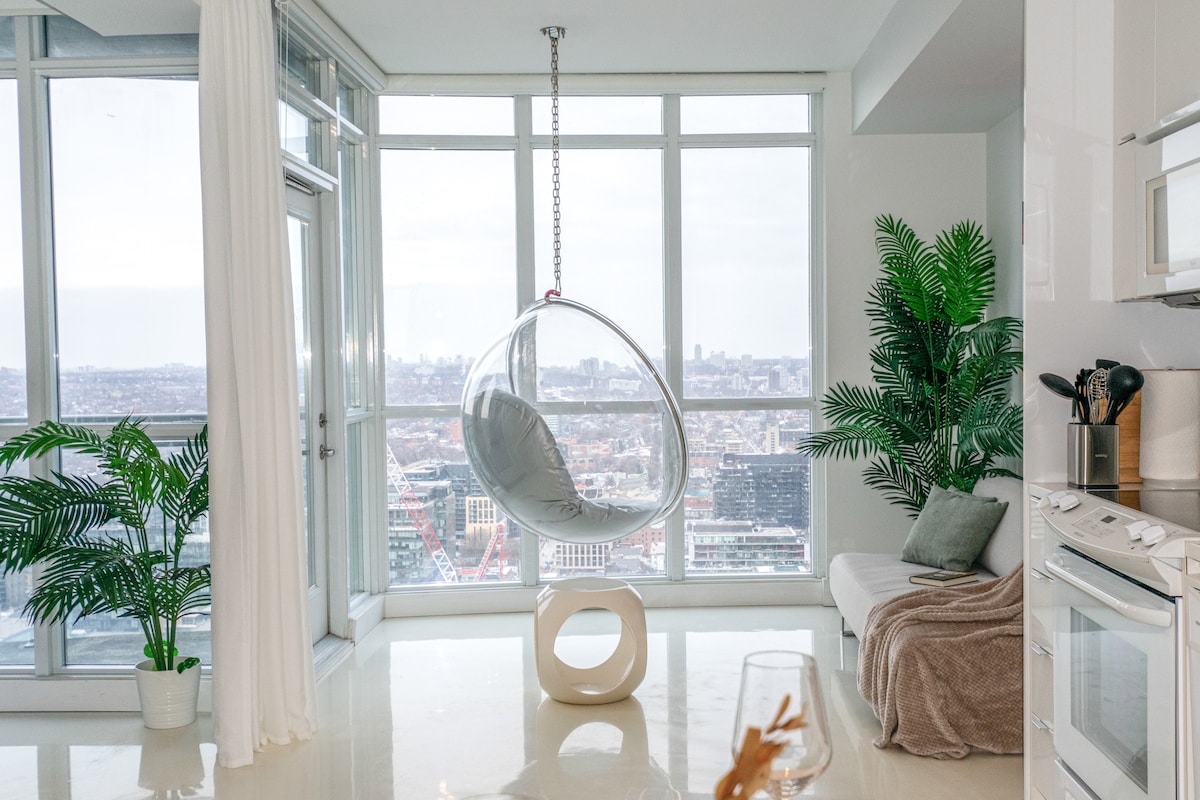 A modern living space is showcased with a distinctive hanging chair positioned by large windows, offering expansive city views. Lush greenery is present, and natural light enhances the room's bright ambiance. A sofa and soft throw complete the cozy seating area.