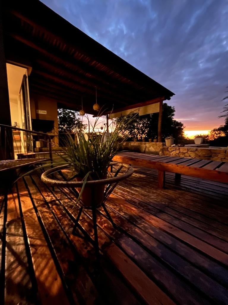 A shaded outdoor deck is illuminated by the soft glow of sunset, featuring wooden lounge seating and a potted plant. The space is designed for relaxation, overlooking natural scenery with a sense of tranquility.