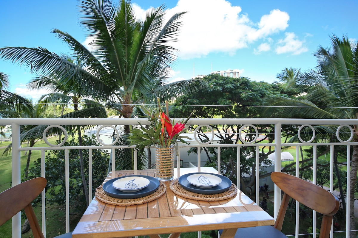 A private lanai features a wooden table set for two, adorned with woven placemats and dishware. Lush palm trees create a tropical ambiance, while bright blue skies provide a serene backdrop. The setting invites relaxation and enjoyment of the surrounding nature.