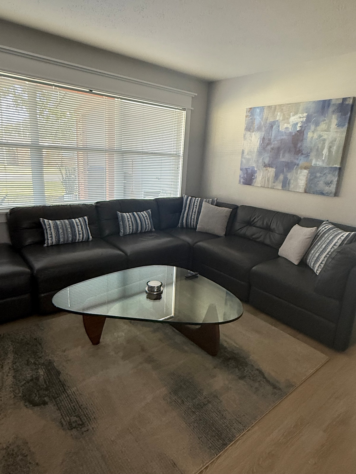 A corner seating area features a dark leather sectional sofa adorned with decorative pillows. A round glass coffee table is positioned in the center, resting on a soft, patterned area rug. Natural light filters through the window, illuminating the space.