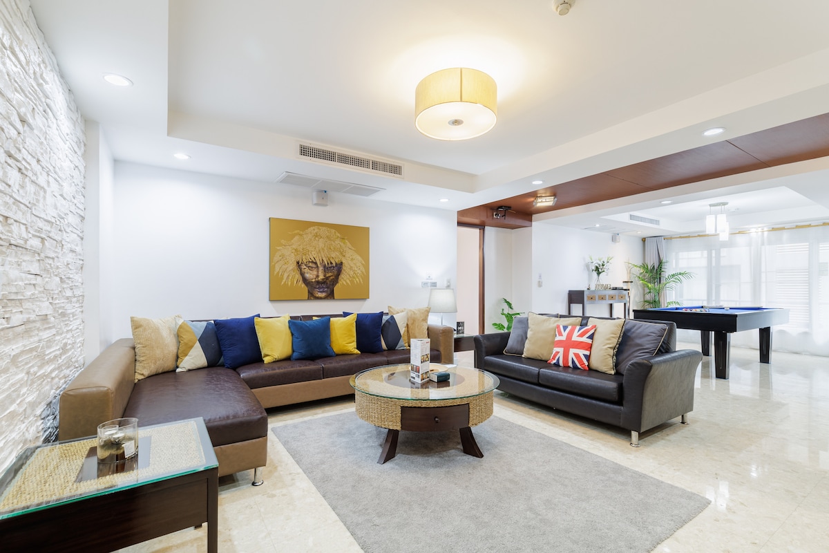 A spacious living area features a combination of modern and comfortable furnishings. A large brown sectional couch and a black leather sofa are complemented by colorful accent pillows. A round glass coffee table rests on a light-colored rug, with soft lighting from the ceiling adding ambiance.