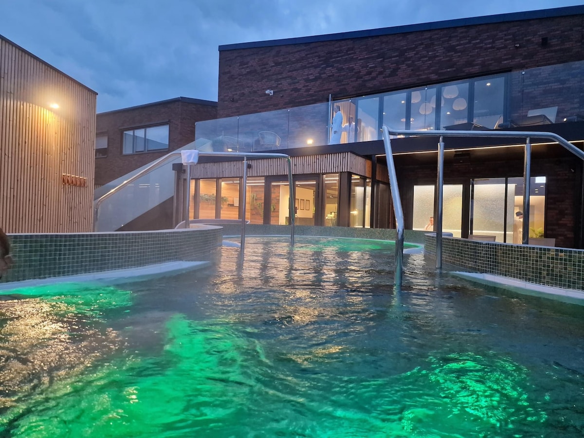 A large heated pool is illuminated in soft green hues, surrounded by contemporary architecture. Glass walls provide a view into the building while creating a seamless transition between indoor and outdoor spaces. Relaxing water features create a tranquil ambiance.