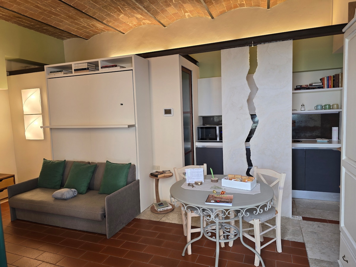 A modern living area features a gray sofa with green cushions, complemented by a round dining table with white chairs. The kitchen area includes a microwave and sleek cabinets, while a decorative wall adds visual interest.
