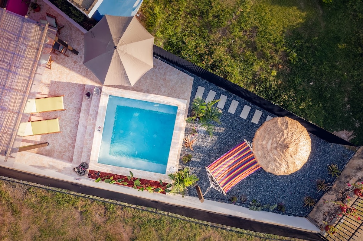 An aerial view showcases a private pool surrounded by a patio area featuring lounge chairs and a large umbrella. A gazebo with vibrant fabric is nearby, complemented by stone pathways and landscaped greenery, providing a serene outdoor space.