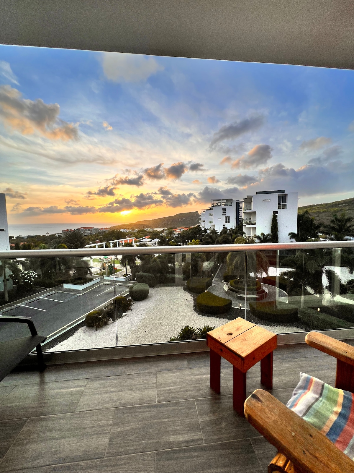 A spacious balcony features wooden chairs overlooking a vibrant sunset. The sky is filled with hues of orange and purple, while manicured gardens and modern buildings are visible in the foreground, creating a tranquil atmosphere.