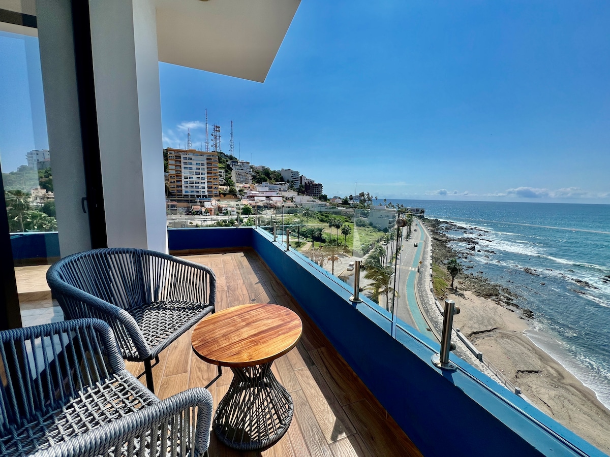 A private balcony is furnished with two woven chairs and a round wooden table. Panoramic views of the ocean and shoreline are visible, with the coastal road winding along the beach. Bright sunlight enhances the serene atmosphere, complemented by clear blue skies.