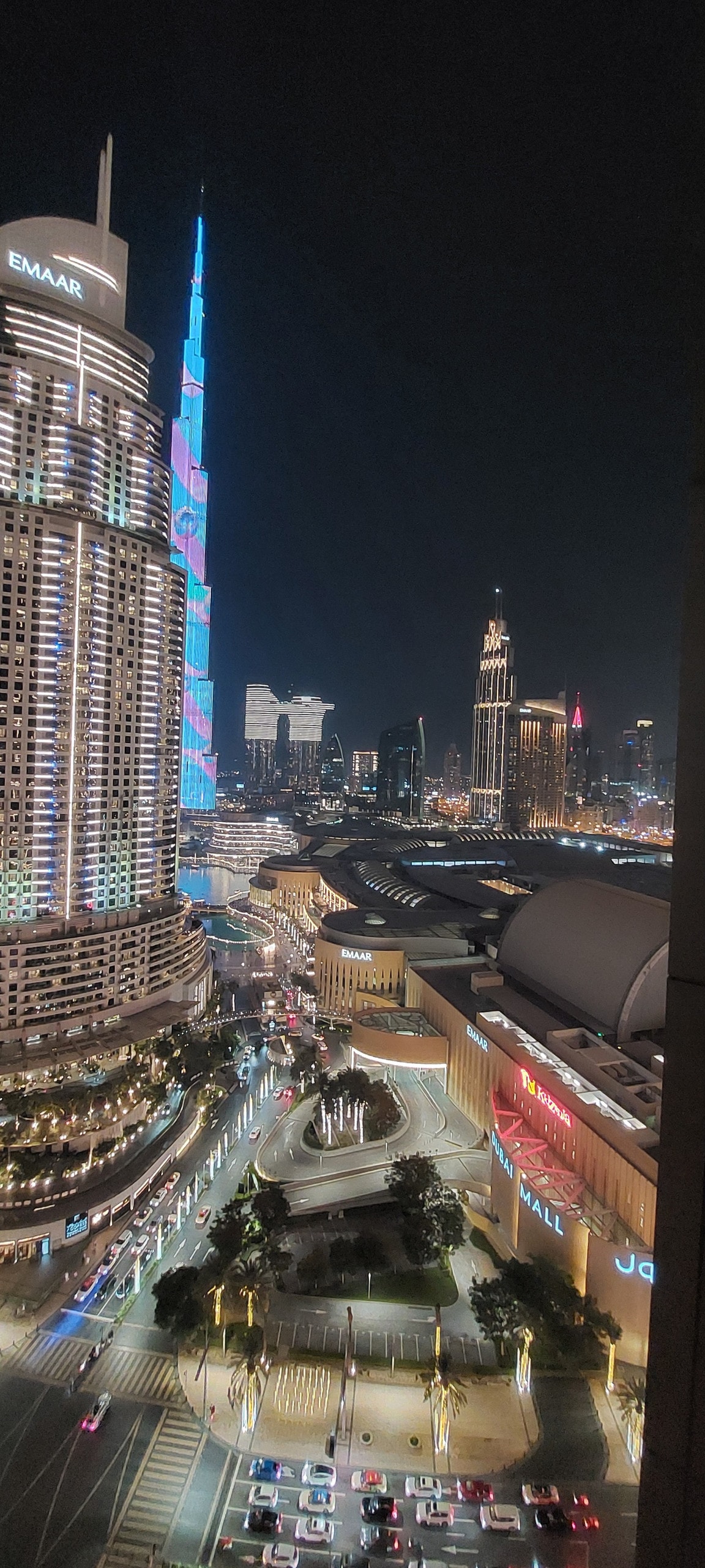 A nighttime view captures the iconic Burj Khalifa and the surrounding skyline, illuminated by city lights. Nearby, the Dubai Mall and surrounding streets are visible, showcasing the dynamic atmosphere of urban life. The scene reflects a blend of modern architecture and vibrant energy.