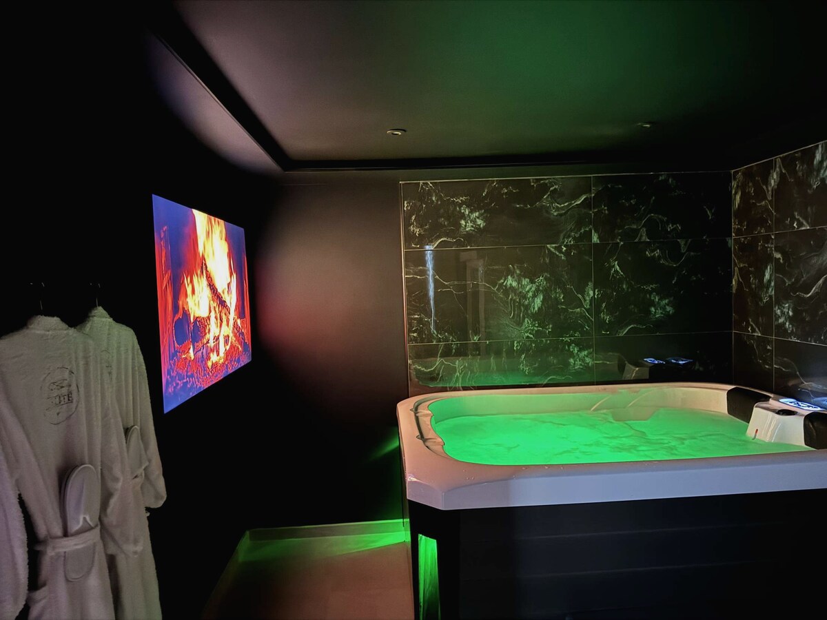 A luxurious private jacuzzi is featured in a dimly lit room, surrounded by sleek black walls and marble accents. A calming green light illuminates the water, while a digital screen displays a flickering fireplace, enhancing the relaxing ambiance. Soft robes are hung nearby for convenience.