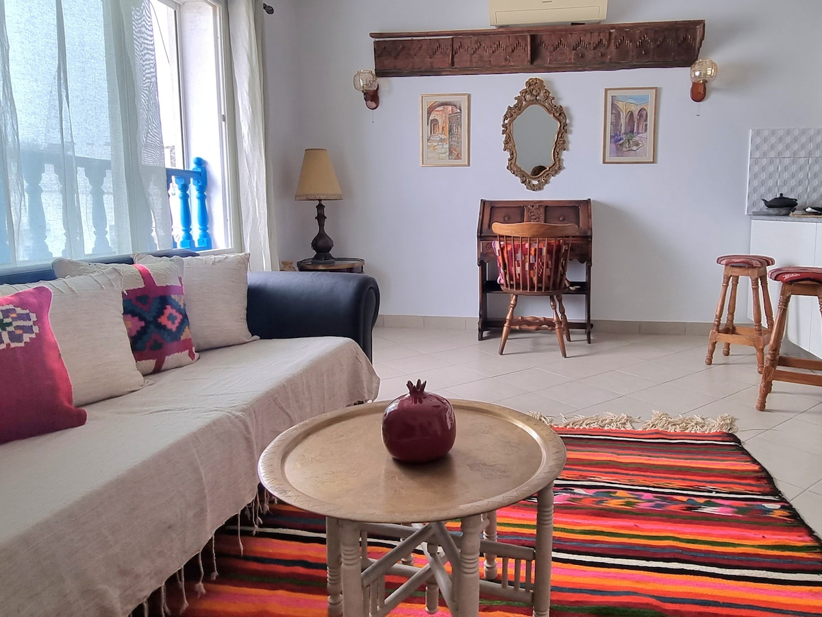 A cozy living area showcases a dark sofa adorned with colorful cushions, complemented by a traditional woven rug. A wooden table sits in the center, featuring a decorative item. A desk with chairs and framed wall art enhances the space, alongside a lamp illuminating the ambiance.