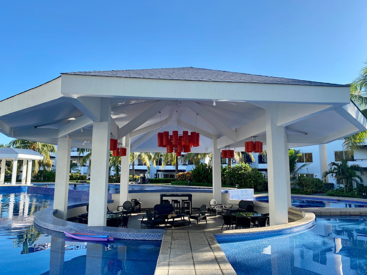 A covered pavilion encircled by water features a dark dining set under red pendant lights. Surrounding palm trees and buildings create a tropical ambiance, and the sun reflects off the calm water, enhancing the serene environment.