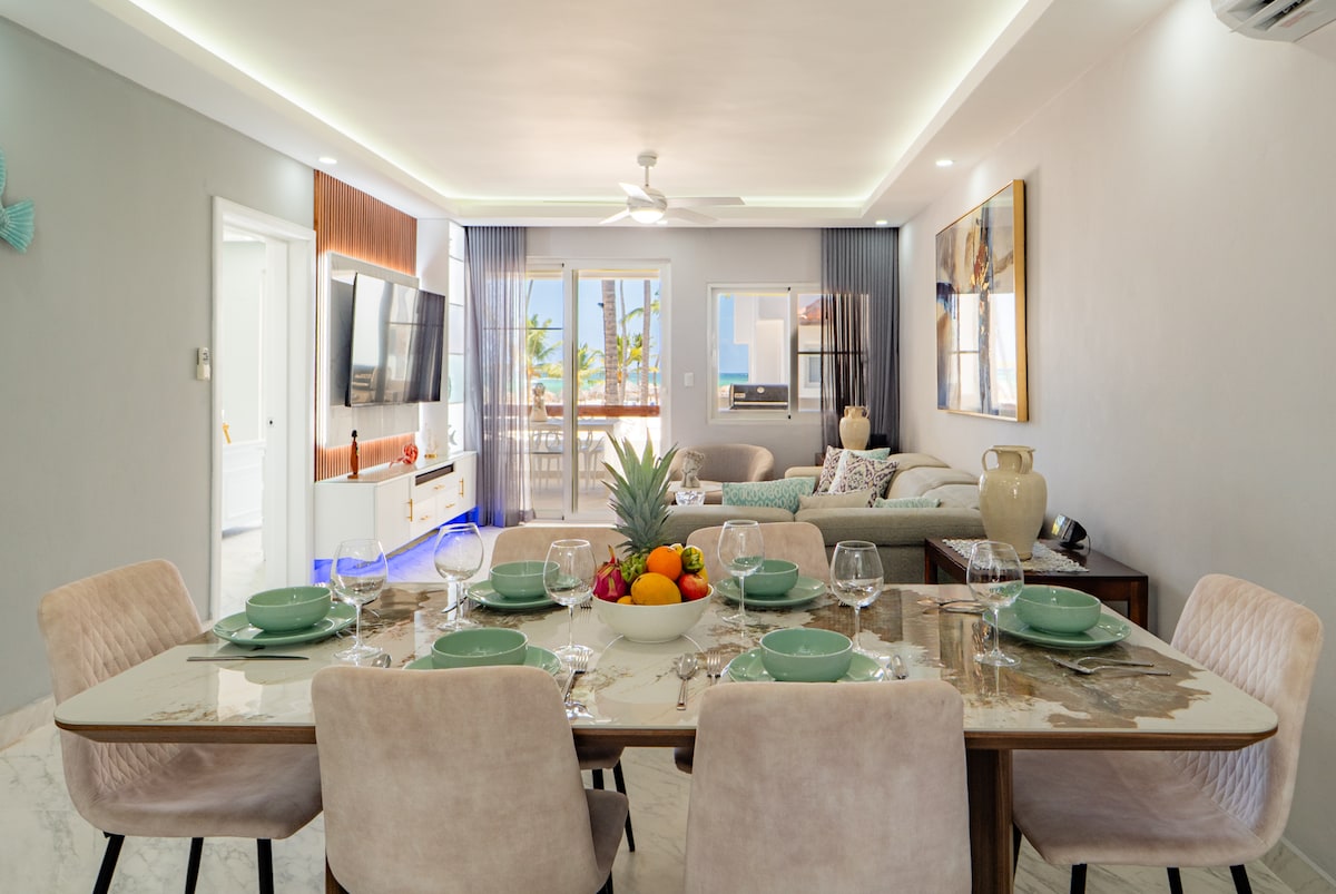 A bright dining area features a table elegantly set with plates and glassware, surrounded by six soft chairs. A large bowl of fresh fruit sits at the center. In the background, a comfortable seating area is visible, along with large windows showcasing views of the ocean.