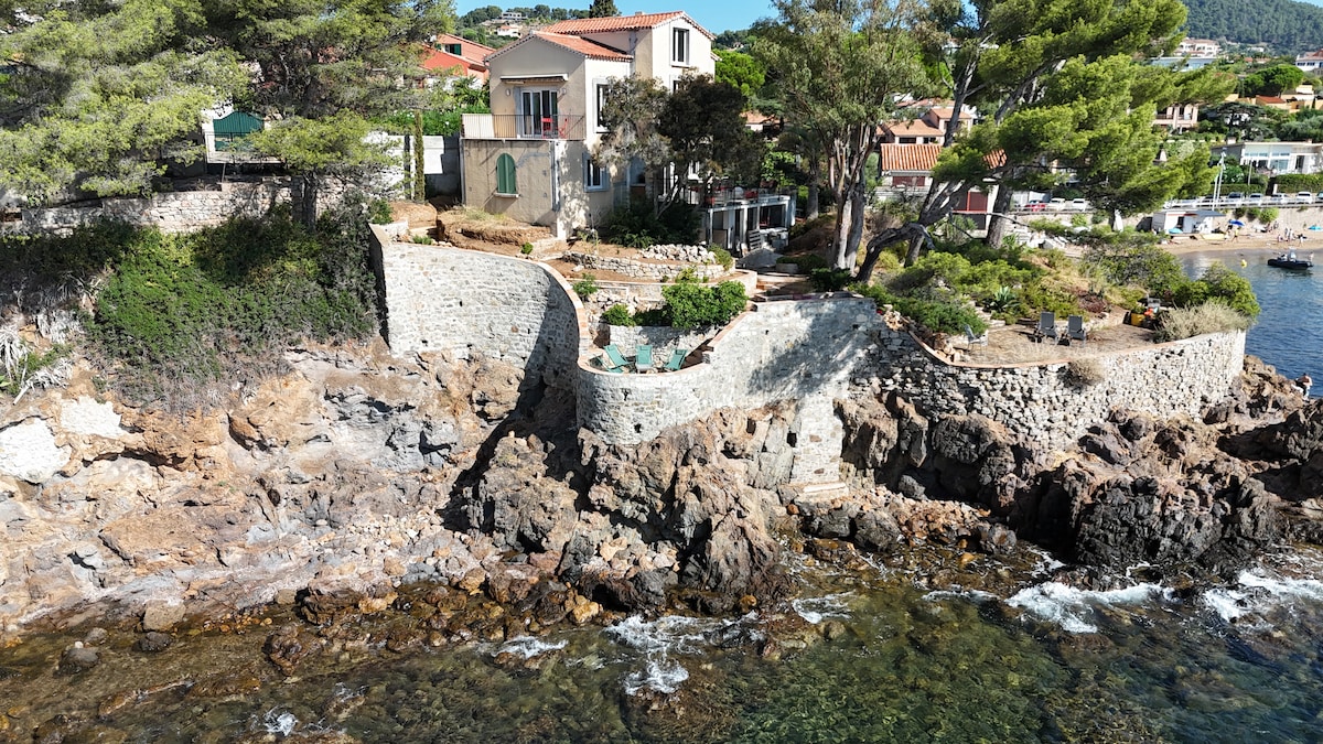 The house is positioned on a rocky shoreline, surrounded by vibrant greenery and the crystal-clear waters of the Mediterranean. A well-maintained garden is visible, along with stone terraces that create inviting outdoor spaces for relaxation and enjoyment.