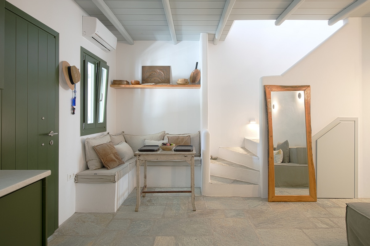 A bright and airy living area features a cozy built-in seating nook beside a rustic wooden table, complemented by decorative bowls. A large mirror reflects light, while a staircase leads to the upper level. Soft colors and natural materials create a calming space.