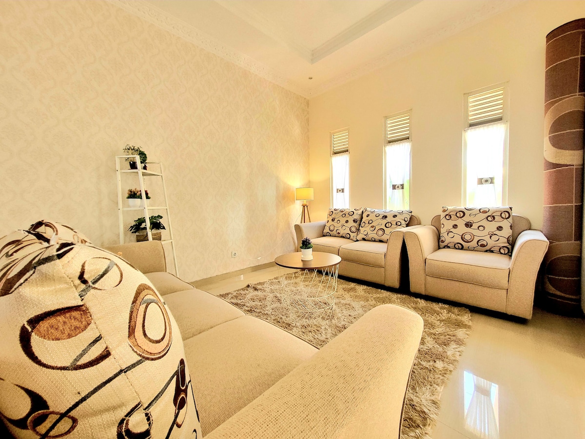 A light-filled living area features beige sofas adorned with circular patterns, set upon a soft area rug. Natural light enters through three windows, enhancing the neutral wall decor. A small coffee table rests in the center, accompanied by a tall plant stand in the corner.