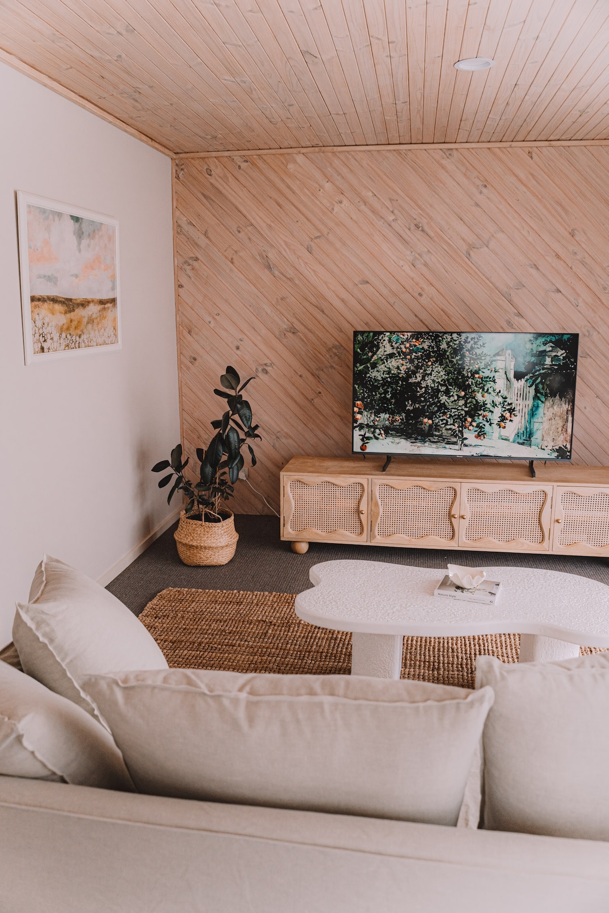 A living area is showcased with light-colored furnishings, featuring a spacious couch and a coffee table. A large TV is positioned against a textured wall, accompanied by a decorative plant in a woven basket. A piece of wall art adds a subtle accent.