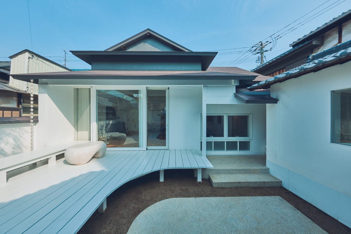 The exterior of the building is characterized by a white cladding and a sloped roof, enhancing its contemporary design. A curved wooden deck leads to large glass doors, inviting natural light into the interior. The calm outdoor space features a simple stone centerpiece.