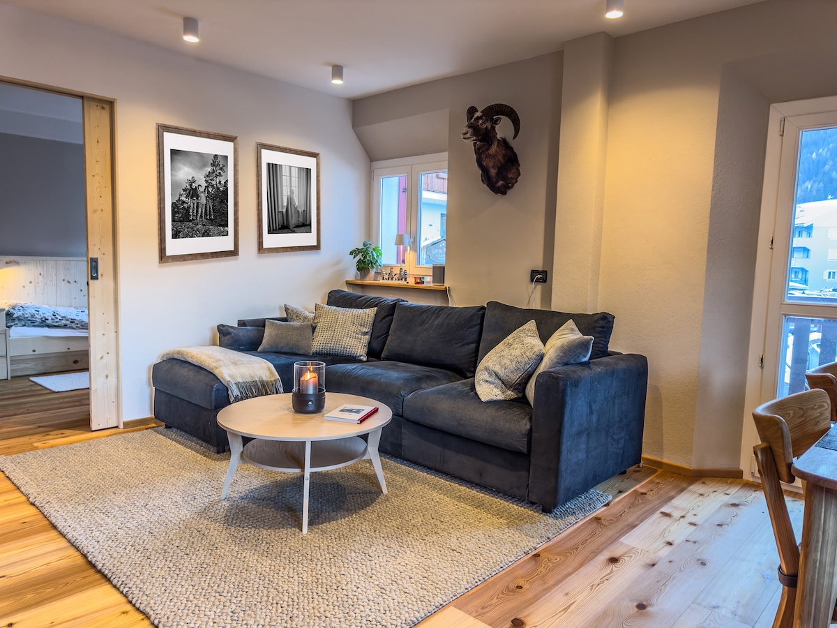 A cozy living area is furnished with a dark blue sectional sofa, accompanied by a round coffee table. Warm wood flooring contrasts with the light gray walls, where framed artworks are displayed. Natural light enters through windows, enhancing the inviting atmosphere.