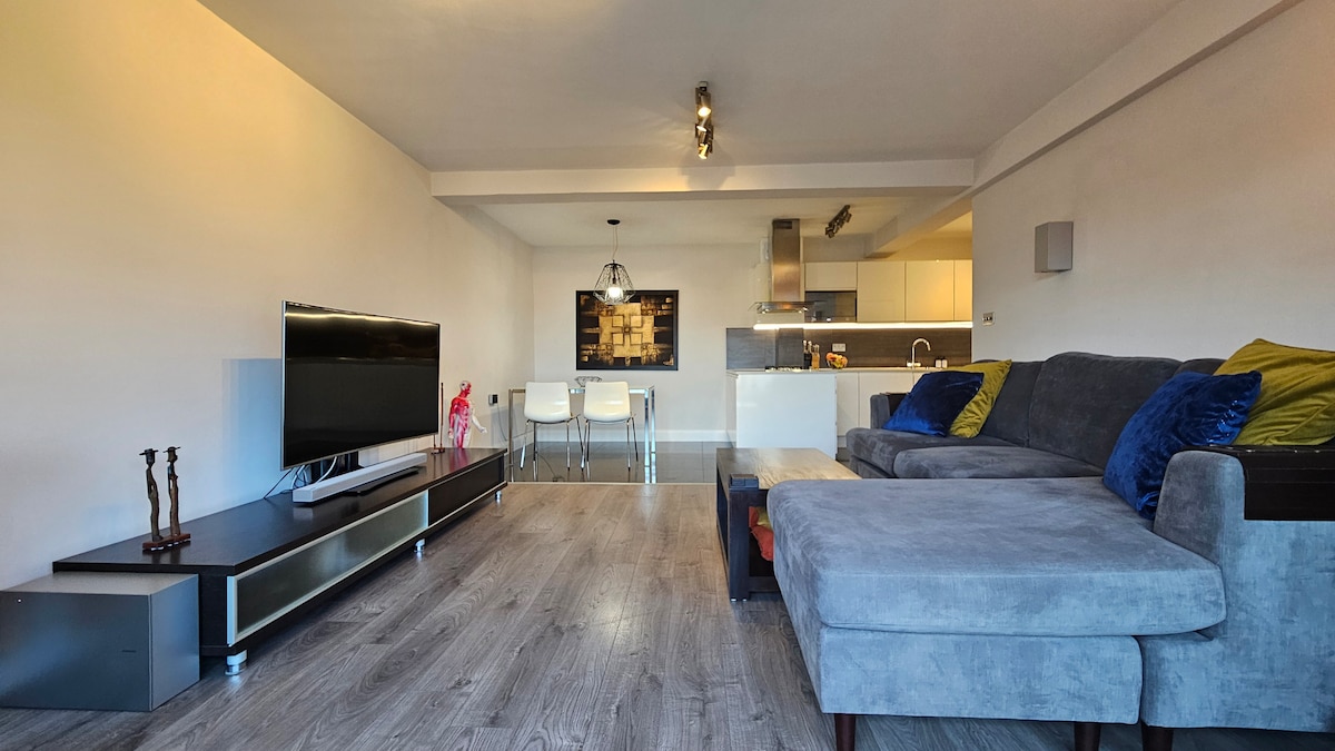 An open living area reveals a spacious lounge with a large sectional sofa and a coffee table. A 50-inch Smart TV is mounted on a media console, and dining seating for four is visible near a modern kitchen featuring sleek appliances.