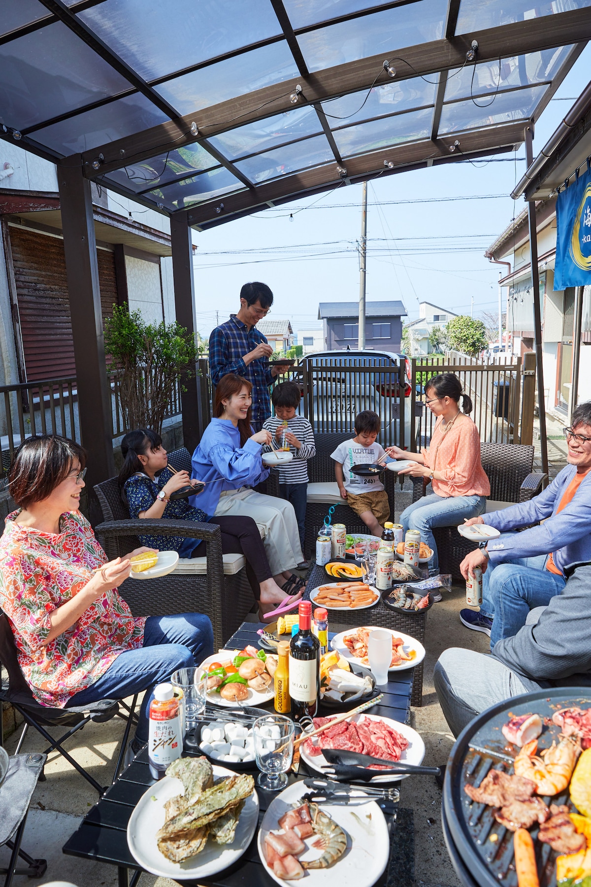 An outdoor dining area features a large table covered with various dishes, including grilled meats and seafood. Guests are seated in comfortable chairs under a protective canopy, enjoying a relaxed meal together. The surrounding space is airy, with natural light enhancing the setting.