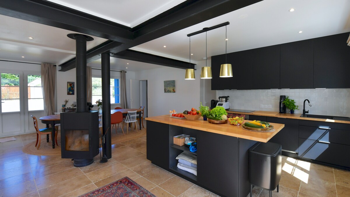 A spacious living area incorporates a dining space, kitchen, and lounge, with a central wood stove providing warmth. An island counter displays fresh produce, flanked by two pendant lights. Large glass doors lead to an outdoor area, enhancing natural light within.