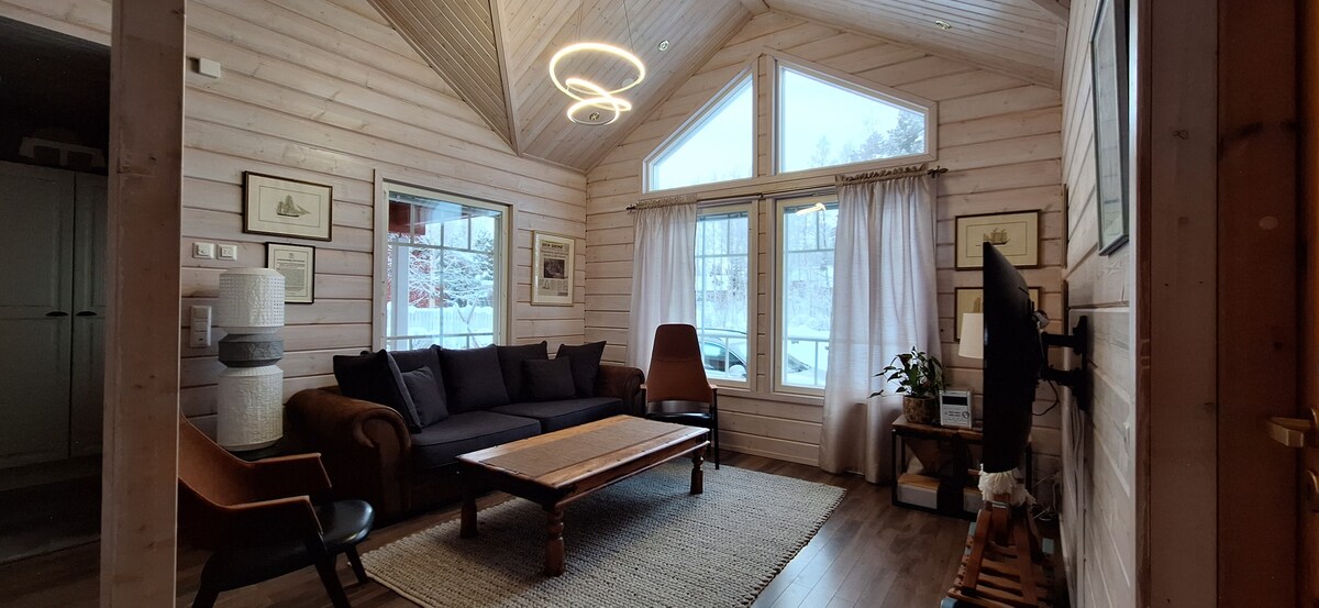 A welcoming living area features large windows that allow natural light to fill the space. A comfortable sofa is positioned across from a modern television stand. A wooden coffee table sits on a textured rug, complemented by stylish armchairs and decorative wall art.