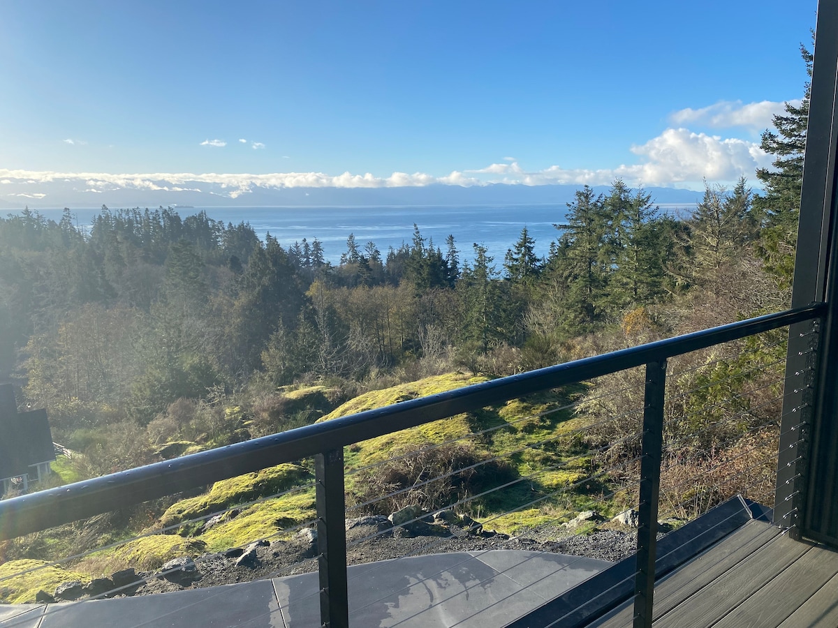 The image showcases an expansive view from the upper balcony, featuring a clear blue sky and the ocean in the distance. Lush greenery and trees are visible in the foreground, blending with the natural landscape. A railing frames the balcony, adding a sense of space.