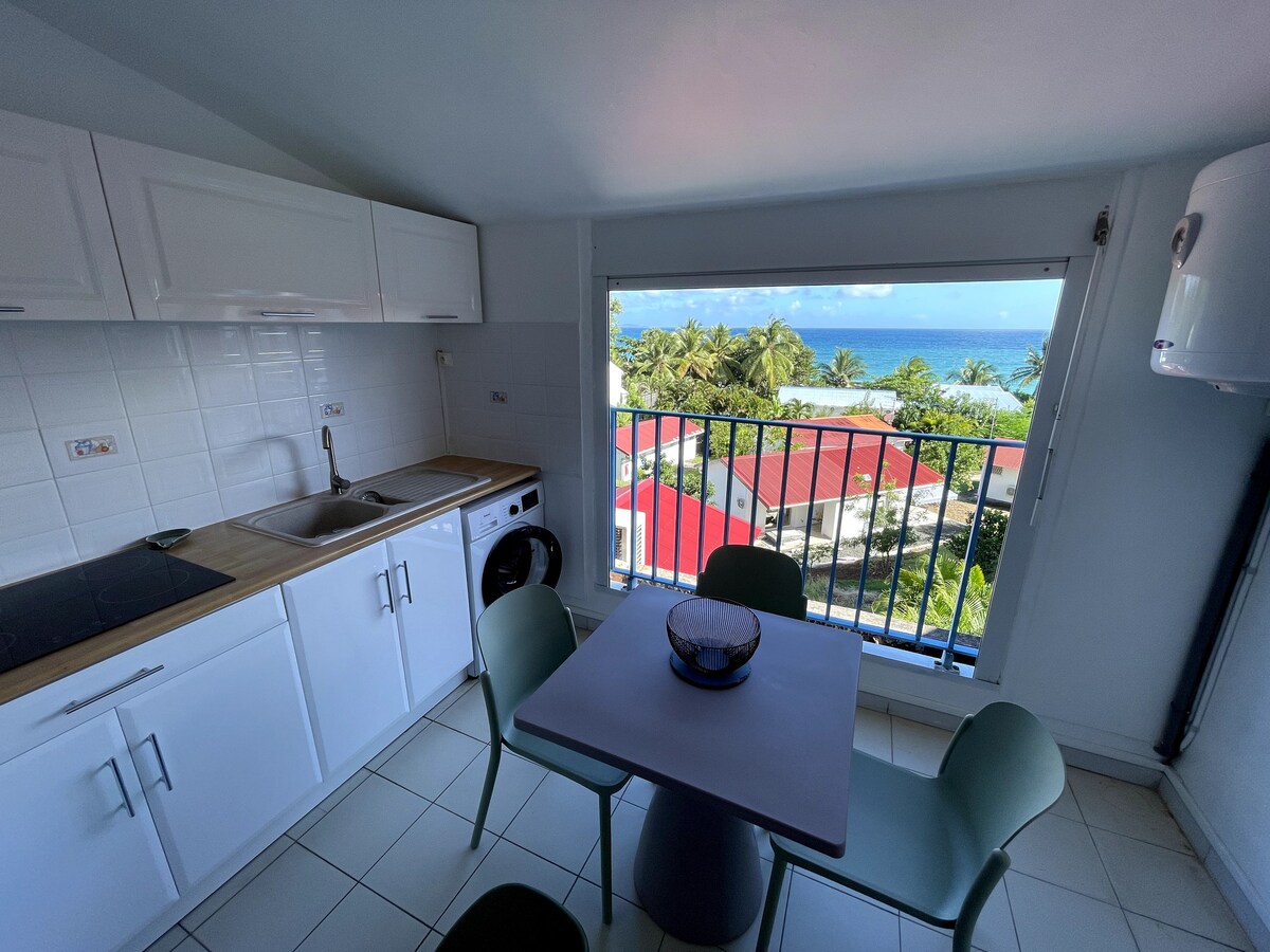 A bright kitchen area is visible, featuring modern cabinetry and essential appliances. A small dining table with green chairs is positioned by a large window, which offers a view of the sea and surrounding landscape, enhancing the sense of openness.