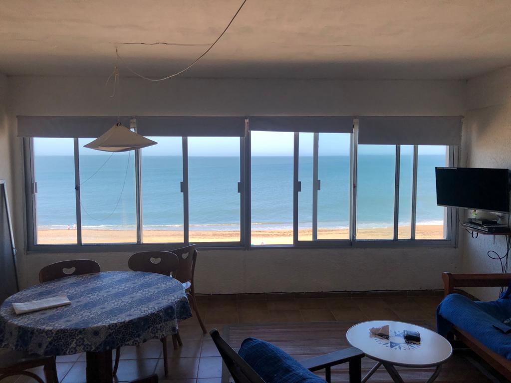 A spacious living area features large sliding windows that offer an expansive view of the beach and ocean. A round dining table is set with a blue-patterned tablecloth, while a small TV is mounted on the wall and a comfortable futon is placed nearby.