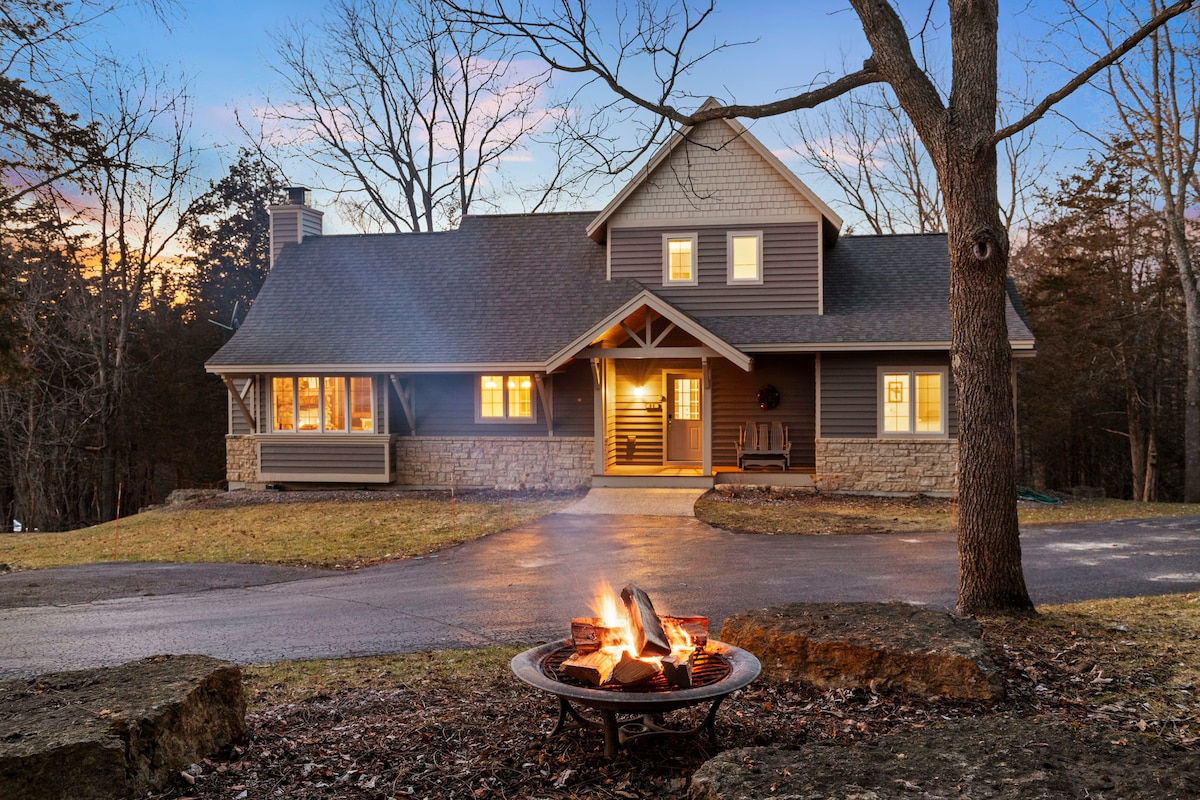The lodge is showcased from outside, highlighting a cozy fire pit surrounded by large stones. Soft light emanates from the windows, with trees framing the picturesque exterior. A paved driveway leads to the entrance, inviting guests to explore the warm ambiance within.