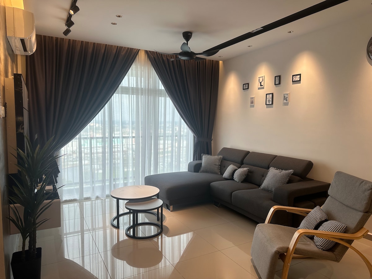 A modern living area features a sectional sofa placed beside a light wood accent chair. A coffee table sits in front, accompanied by a floor plant. Natural light filters through sheer curtains, highlighting framed pictures on the wall and creating a spacious feel.