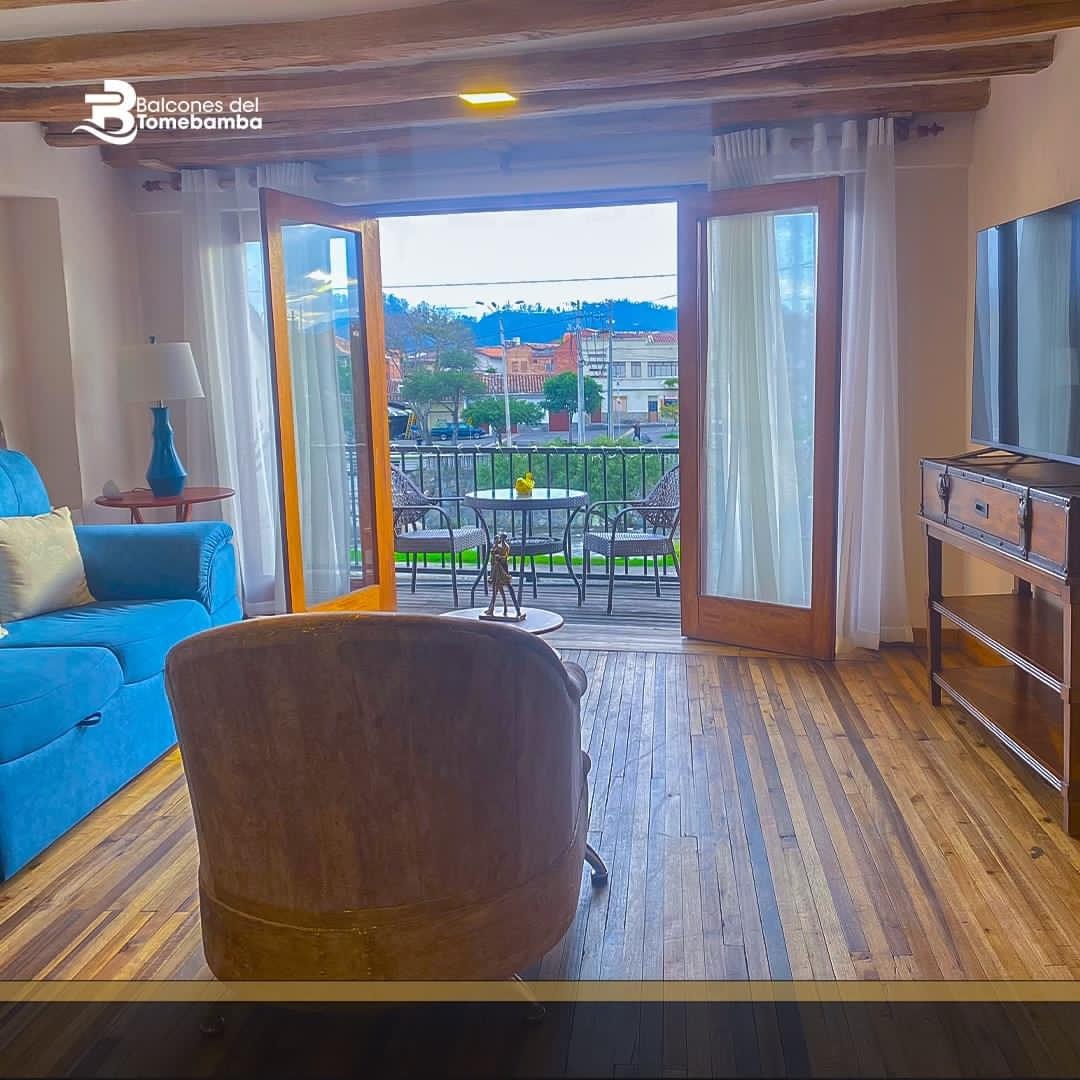 A comfortable living area features a blue sofa, wooden accents, and a coffee table. Large glass doors open to a balcony with outdoor seating, providing a view of the surrounding landscape. The floor is composed of warm wood, enhancing the inviting atmosphere.
